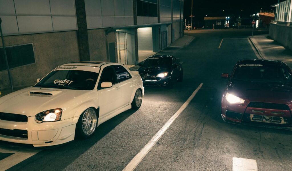 Three modified sports cars a white Subaru Impreza, a black Subaru BRZ, and a red Mitsubishi Lancer Evolution — are parked at night on an urban street, illuminated by streetlights.