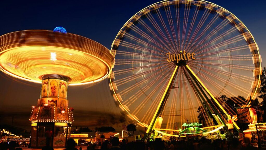 A brightly lit amusement park at dusk featuring a large spinning swing ride on the left and a glowing Ferris wheel labeled "Jupiter" on the right, with crowds of people enjoying the festive atmosphere below.