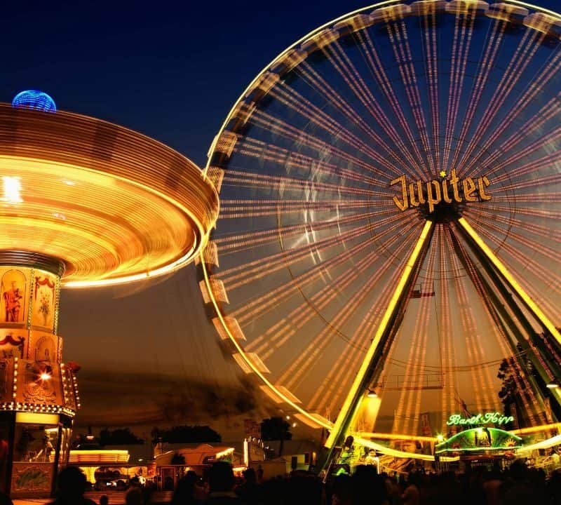 A brightly lit amusement park at dusk featuring a large spinning swing ride on the left and a glowing Ferris wheel labeled "Jupiter" on the right, with crowds of people enjoying the festive atmosphere below.