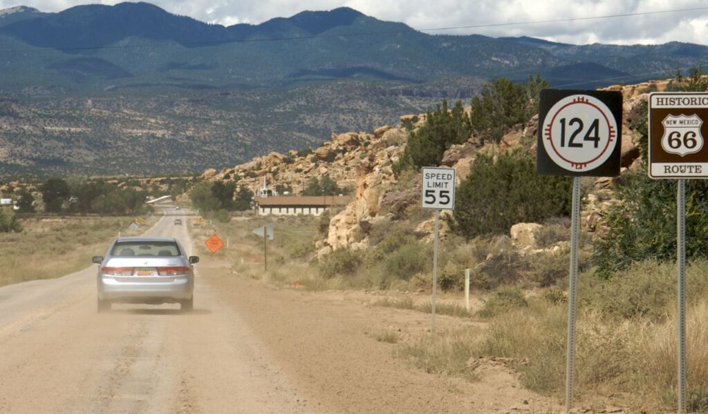 A silver car drives along a dusty stretch of Historic Route 66 in New Mexico, surrounded by desert terrain, road signs, and scenic mountains in the background.