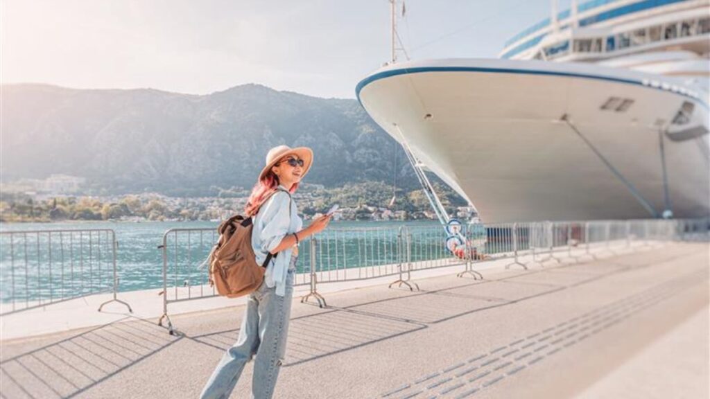 A woman in a sunhat and sunglasses stands near a large cruise ship docked at a scenic port, holding her phone and wearing a backpack, with mountains and water in the background.