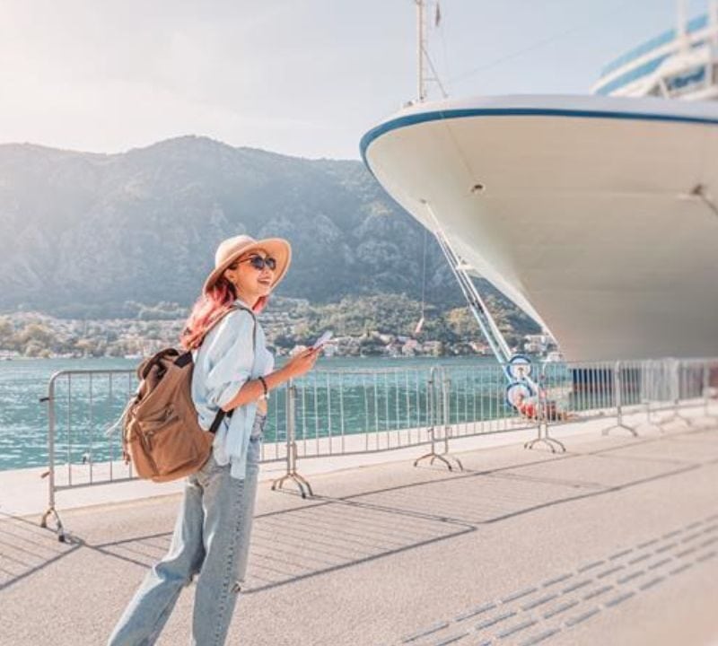A woman in a sunhat and sunglasses stands near a large cruise ship docked at a scenic port, holding her phone and wearing a backpack, with mountains and water in the background.