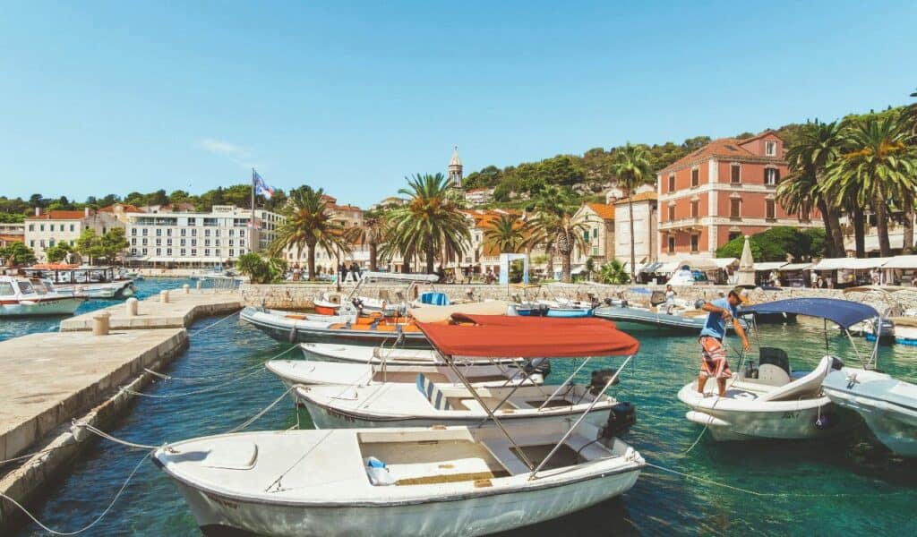 Small boats docked in the turquoise harbor of Hvar, Croatia, with palm trees, historic buildings, and a sunny sky in the background.