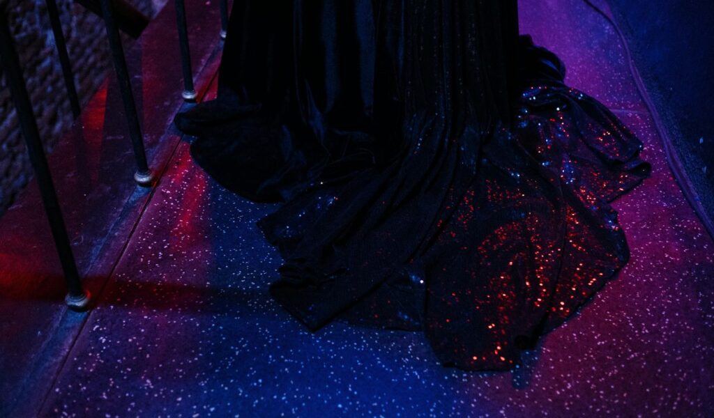The image shows a black gown with sparkling sequins, highlighted by red and purple lighting, creating an elegant, glamorous vibe.