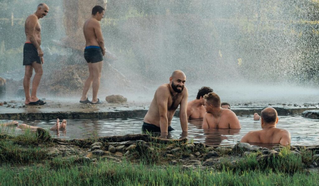 Group of men in swim trunks relaxing in a natural hot spring with steam rising, while others stand under a shower area nearby, surrounded by greenery and mist.
Ask ChatGPT