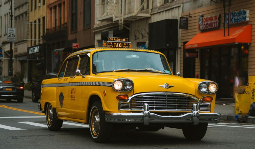 Classic yellow taxi driving through a city street, surrounded by shops and buildings, symbolizing urban transportation.