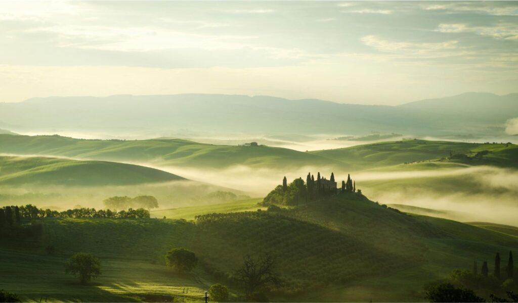 A dreamy, fog-covered landscape of rolling green hills with a cluster of trees and buildings on a hilltop at sunrise.