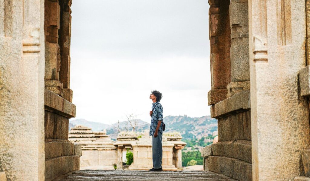 Traveler stands between ancient stone pillars in India, gazing upward, with temple ruins and hills in the background.