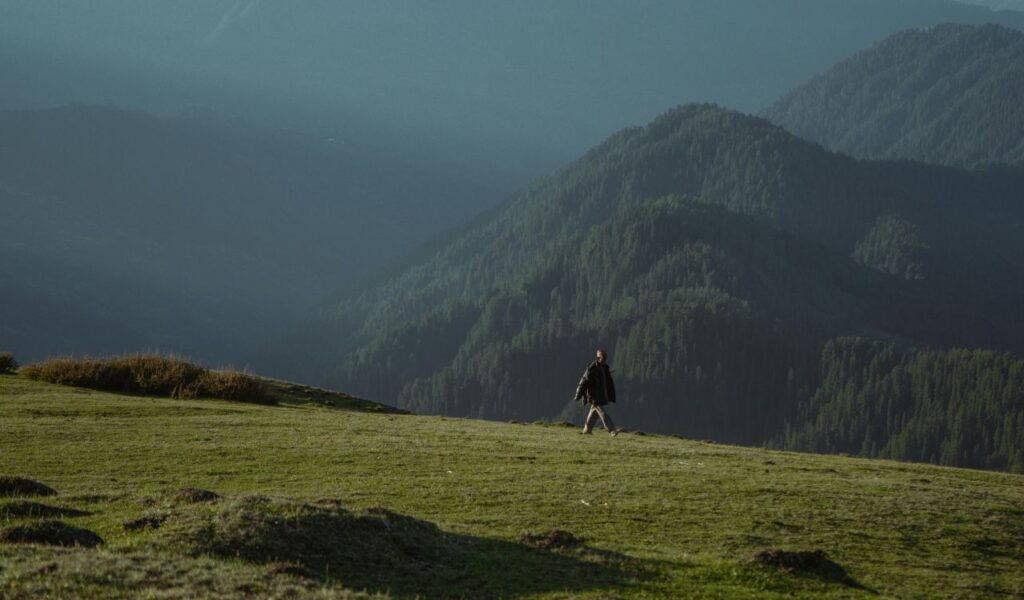 Solo traveler walking on green hillside with forested mountains in the background in India.