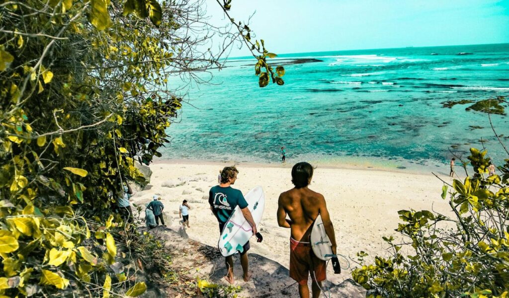 Surfers carrying boards walk down a tropical path toward a sandy beach and turquoise water in Bali, Indonesia.