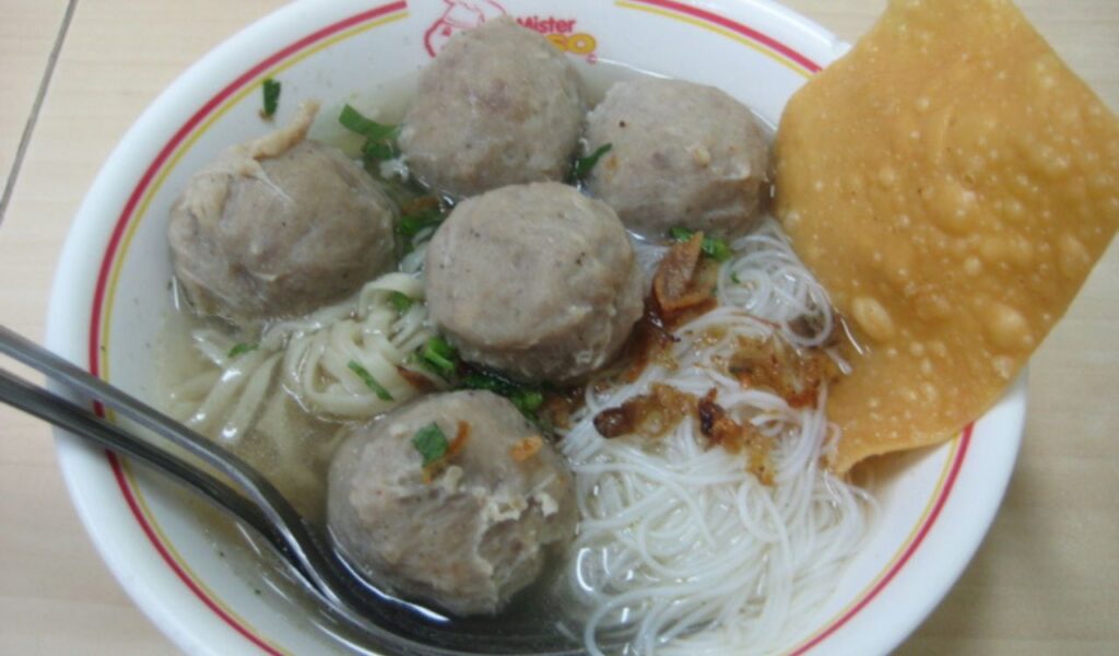 A bowl of Indonesian bakso with beef meatballs, noodles, fried shallots, herbs, clear broth, and a crispy cracker on top.