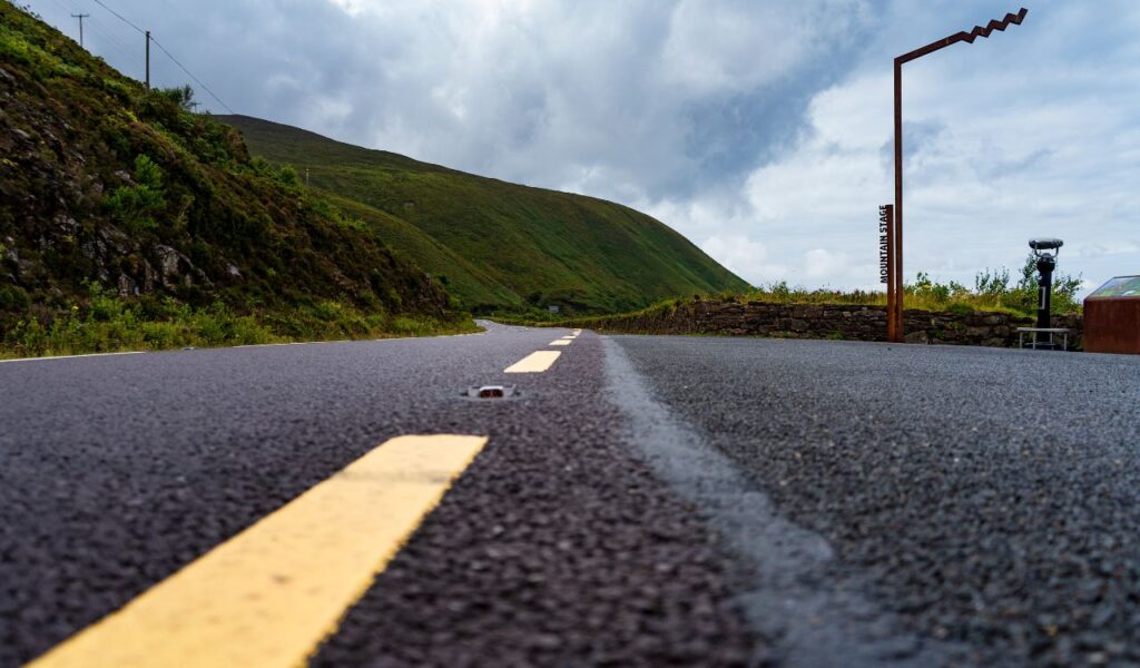 Low-angle view of an empty mountain road with a yellow centerline, surrounded by lush green hills under a cloudy sky. A sign labeled "Mountain Stage" and a scenic viewpoint are visible on the right.