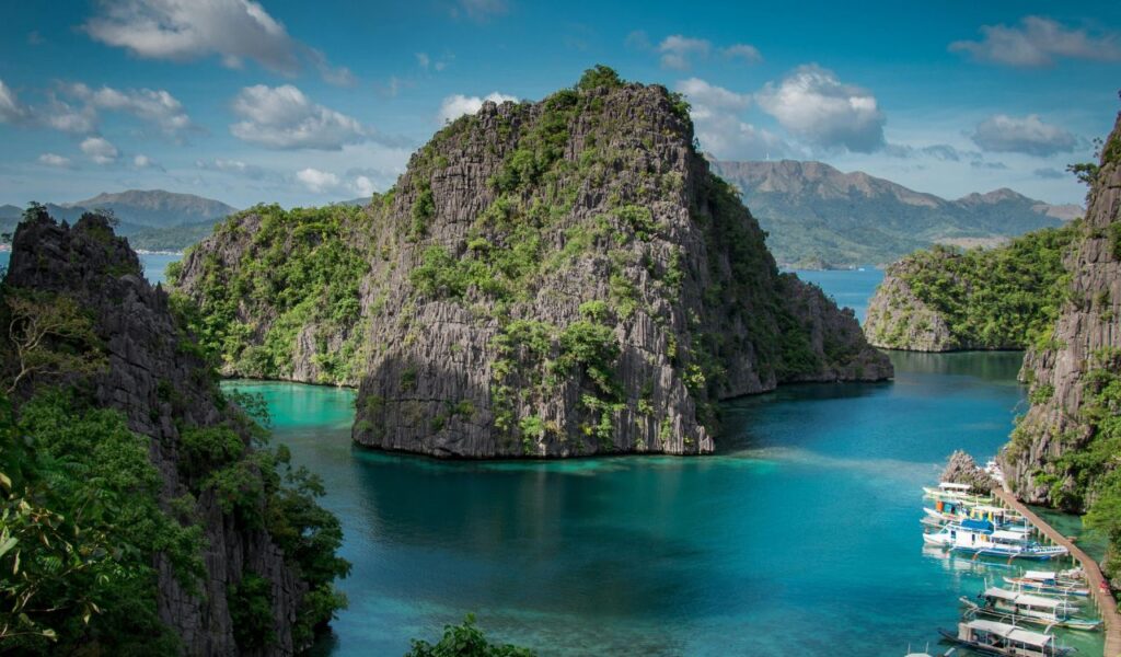 Dramatic limestone cliffs rise from turquoise water, with tour boats docked along a lush tropical island in the Philippines.