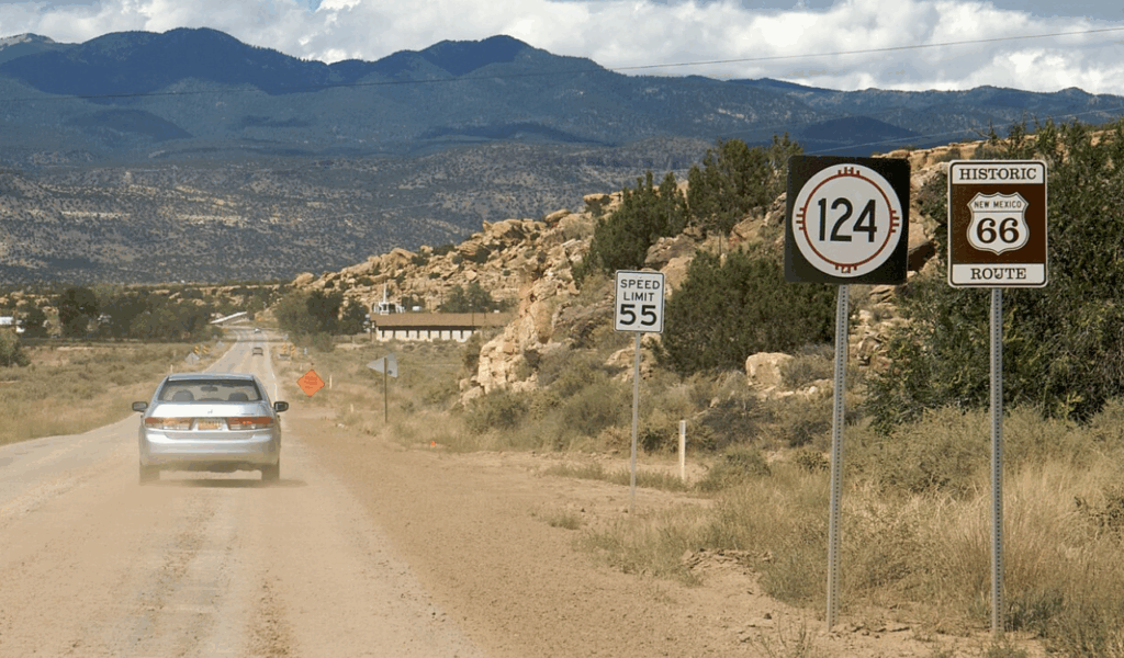 A car drives down a dusty stretch of historic Route 66 in New Mexico, passing speed limit and highway signs, with rugged mountains in the background.