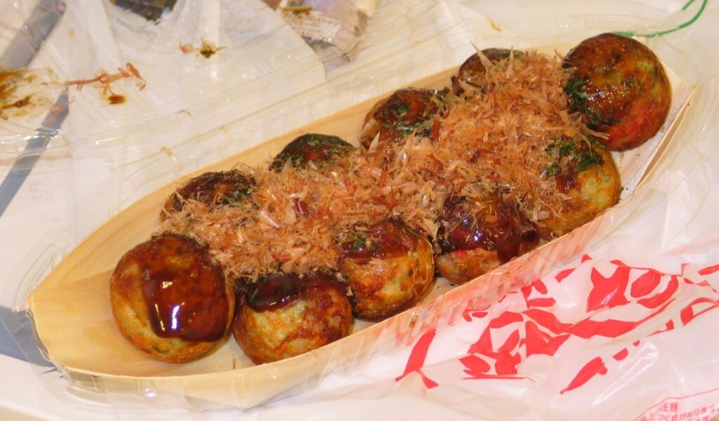 A tray of Japanese takoyaki topped with sauce and bonito flakes, served in a wooden boat-shaped dish with plastic wrapping.