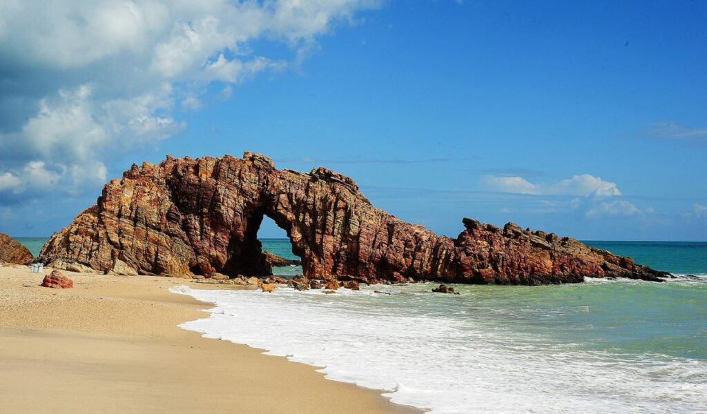 Rock arch at Jericoacoara Beach, Brazil, with clear blue sky and ocean waves.