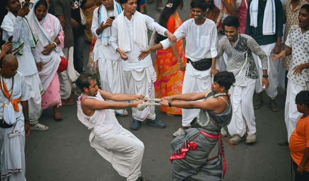  Two men engaged in a traditional tugging ritual during a lively street celebration, surrounded by people in festive Indian attire, clapping and cheering.