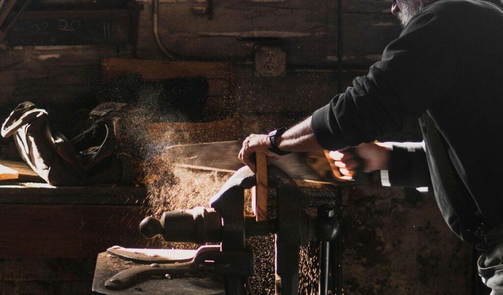 Man using a hand saw to cut wood in a dimly lit workshop, with sawdust flying through the air and tools visible in the background.