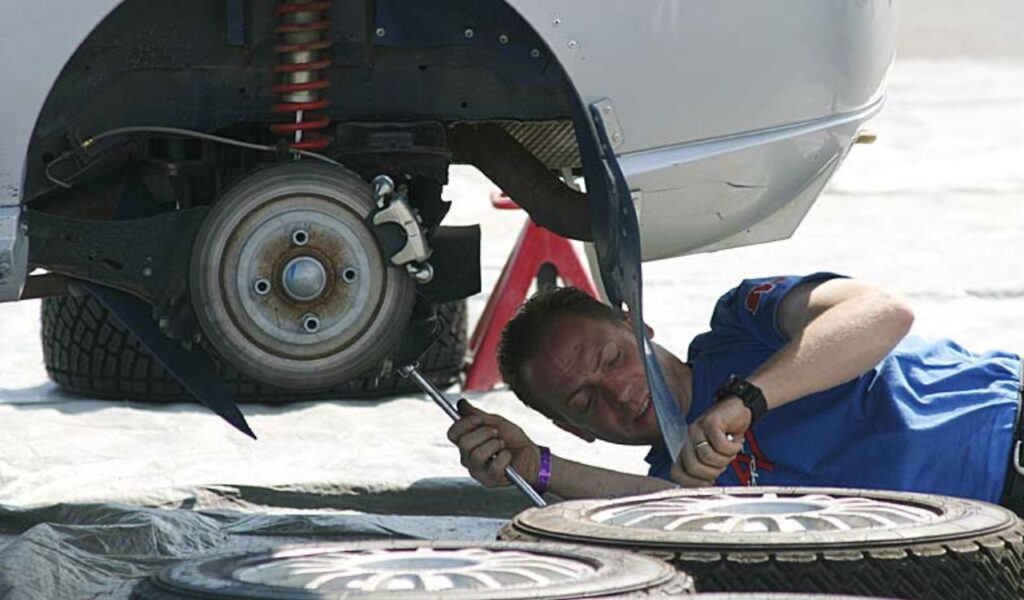 Man working underneath a raised car with removed tires, emphasizing the importance of regular vehicle maintenance.