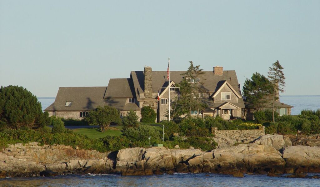 Oceanfront view of the Bush family estate in Kennebunkport, Maine, with rocky shoreline, manicured lawn, and American flag.