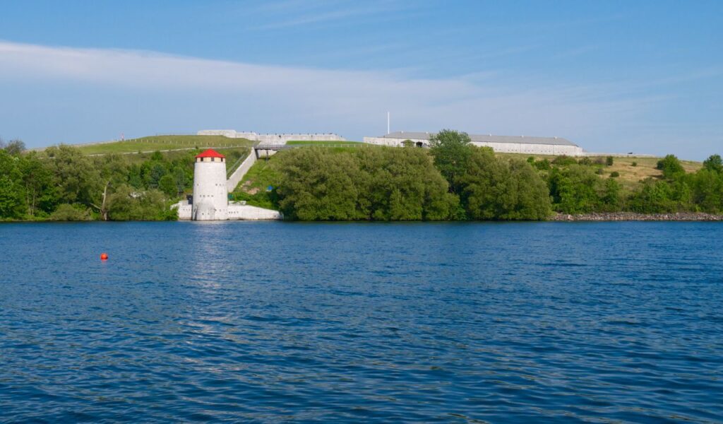 A view across the water of Fort Henry in Kingston, Ontario, with a red-roofed Martello tower surrounded by trees and historic limestone buildings atop the hill.