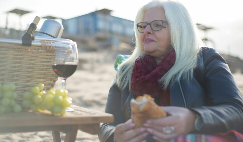 An older woman enjoying a relaxed picnic on the beach, with a glass of wine, grapes, and a sandwich.