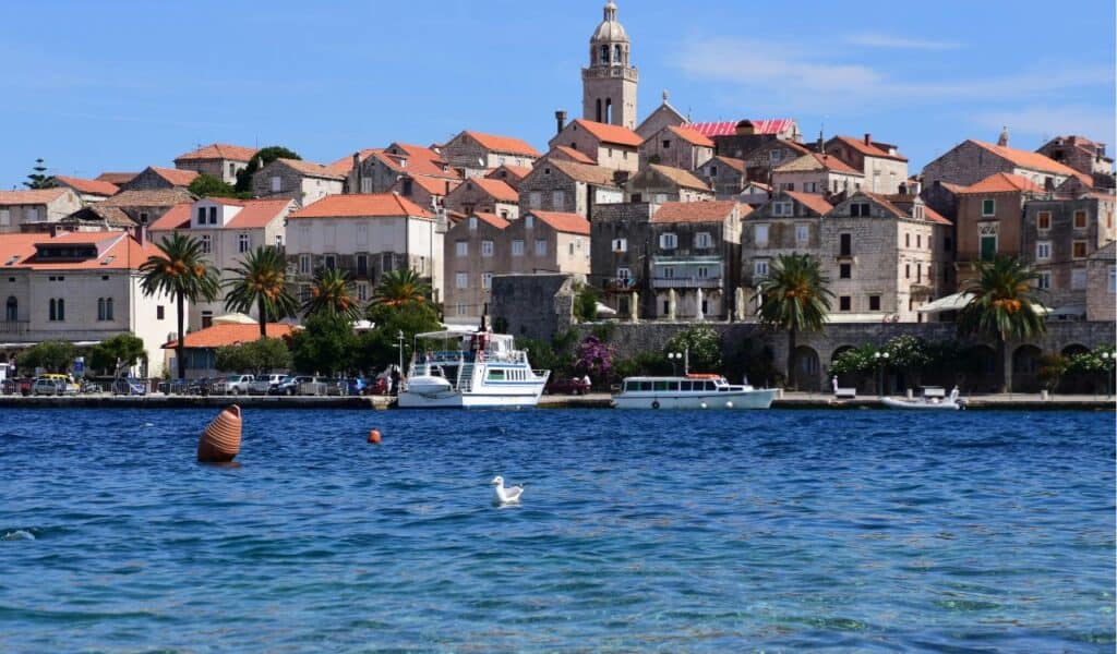 View of Korčula’s old town with stone buildings and red rooftops rising above the Adriatic Sea, boats docked along the waterfront, and a bell tower in the background.