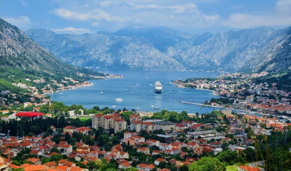 Scenic view of Kotor Bay in Montenegro, framed by rugged mountains, with a cruise ship sailing toward the red-roofed coastal town.