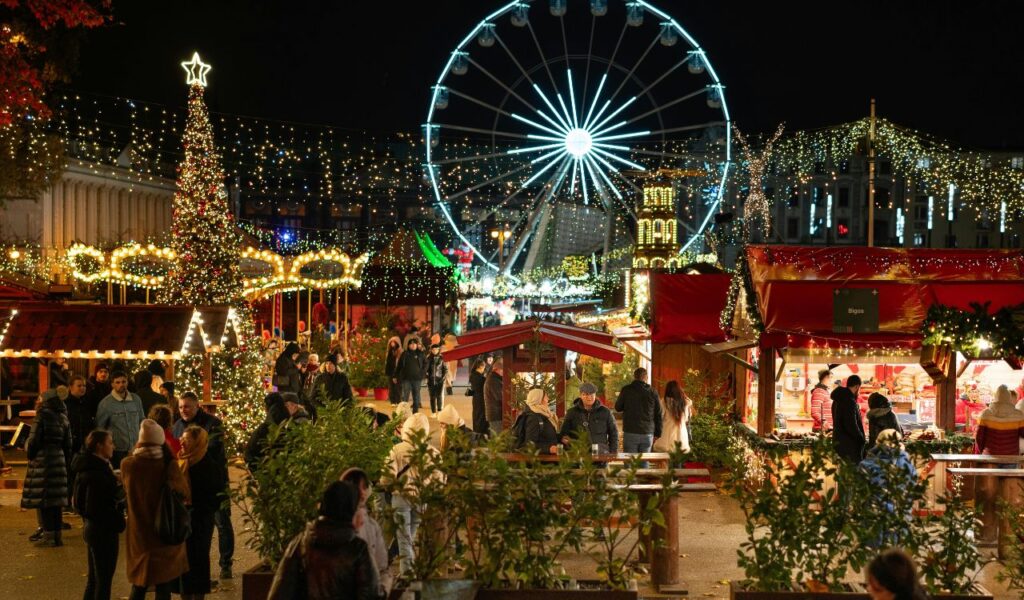 Festive Krakow Christmas Market at night with glowing stalls, a large decorated Christmas tree, sparkling lights overhead, and a bright Ferris wheel in the background.