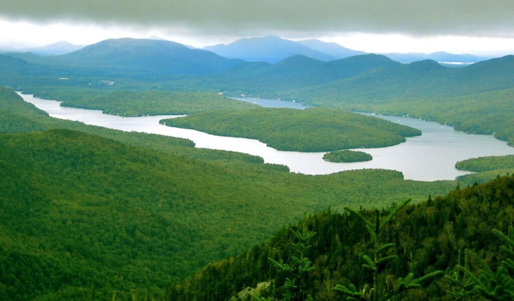 A panoramic view of Lake Placid surrounded by lush green Adirondack Mountains under a cloudy sky in upstate New York.