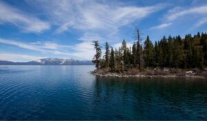 Lake Tahoe shoreline with clear blue water, pine trees, and snow-capped mountains in the distance.