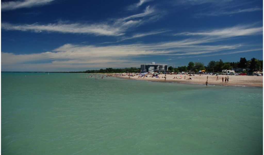 A sunny day at Grand Bend Beach in Lambton Shores, with people relaxing on the sandy shoreline and swimming in the turquoise waters of Lake Huron.
