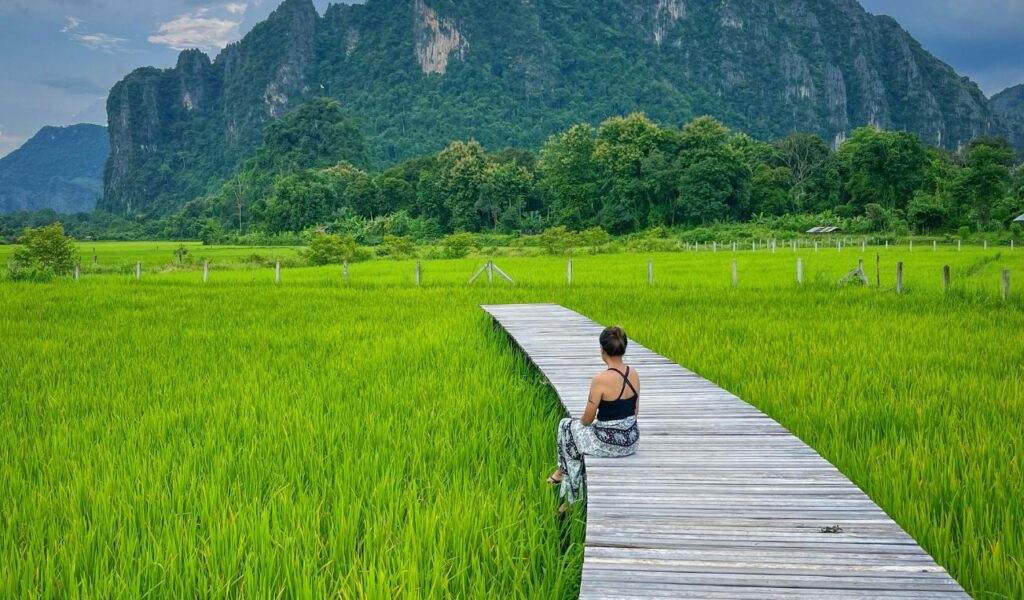 Solo traveler sitting on wooden path through lush rice fields with mountains in Laos.