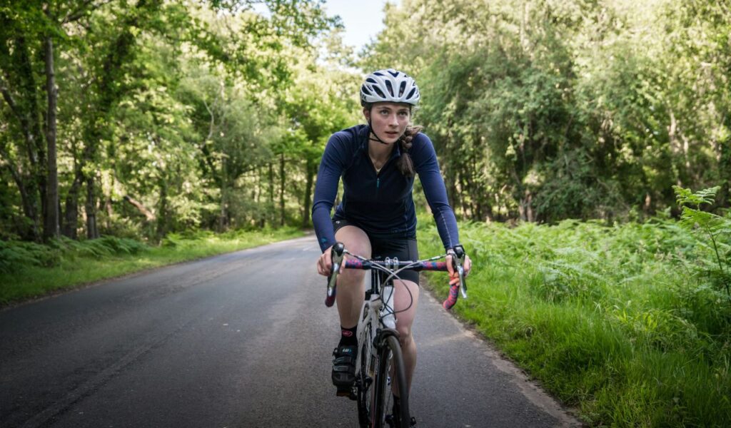 Woman in cycling gear and helmet riding a road bike on a quiet, tree-lined path, focused and determined.