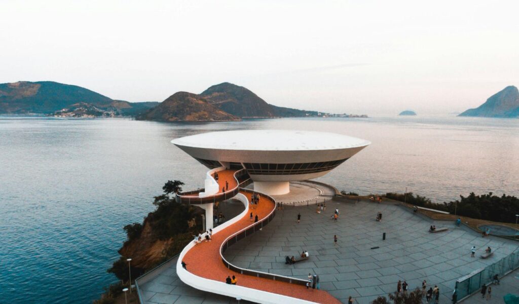 A modern, UFO-shaped building perched on a coastal cliff in Brazil, with a winding walkway leading up to it, surrounded by calm waters and distant mountains at dusk.