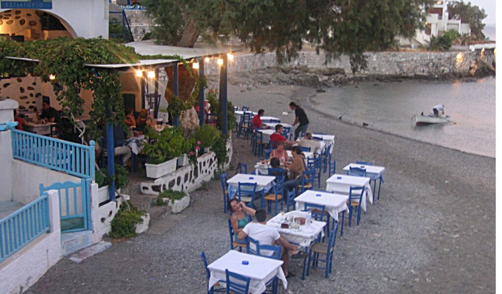 Seaside Greek taverna with white tablecloths and blue chairs set along the shore, where diners enjoy a relaxed evening meal.