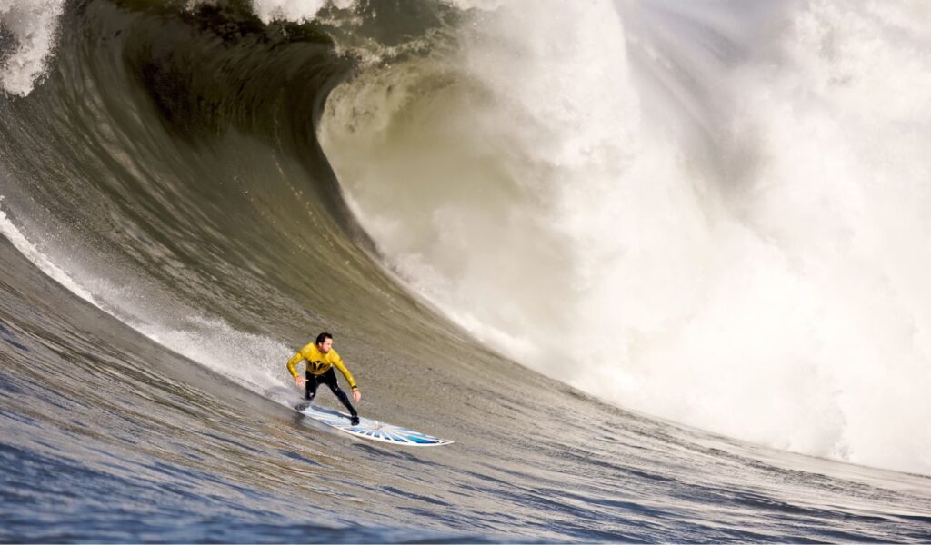 A surfer in a yellow jersey rides a massive wave, highlighting the power and danger of surfing in high-surf locations.