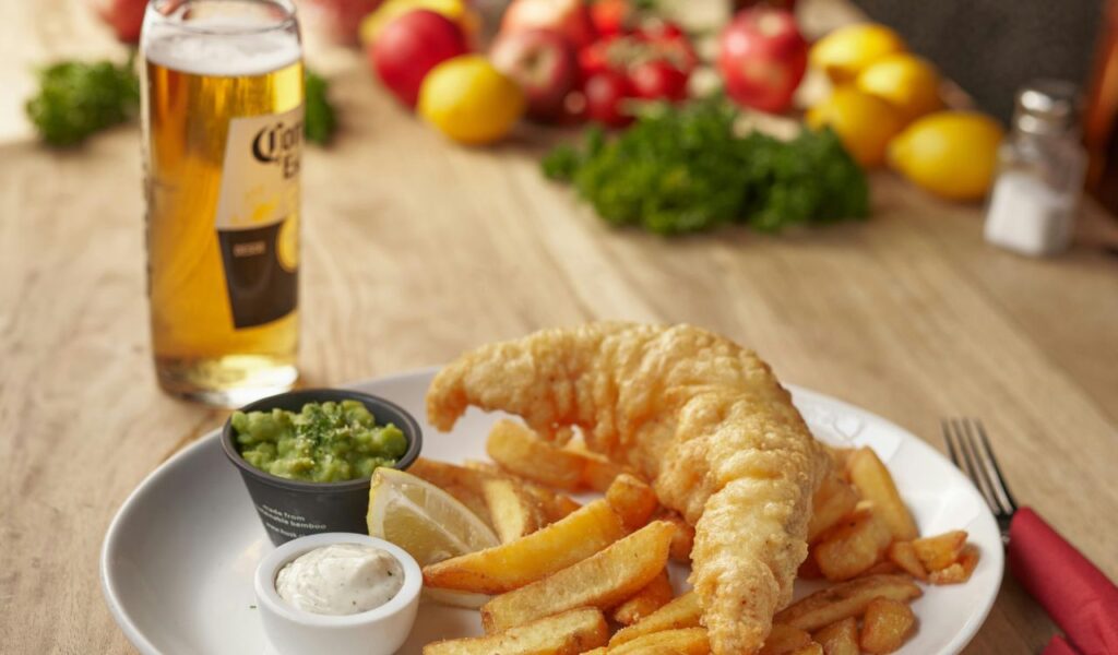 A plate of fried fish with golden fries, mushy peas, lemon wedges, and tartar sauce, served with a glass of Corona beer on a wooden table.