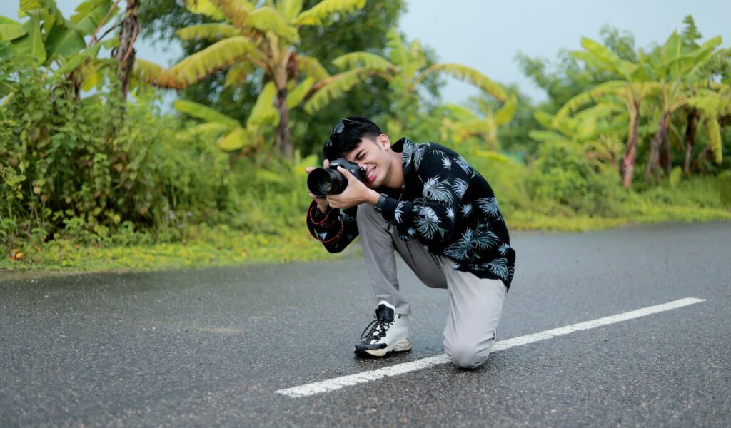 Smiling man kneels on a wet road with lush greenery around, holding a camera and capturing a photo in the moment.