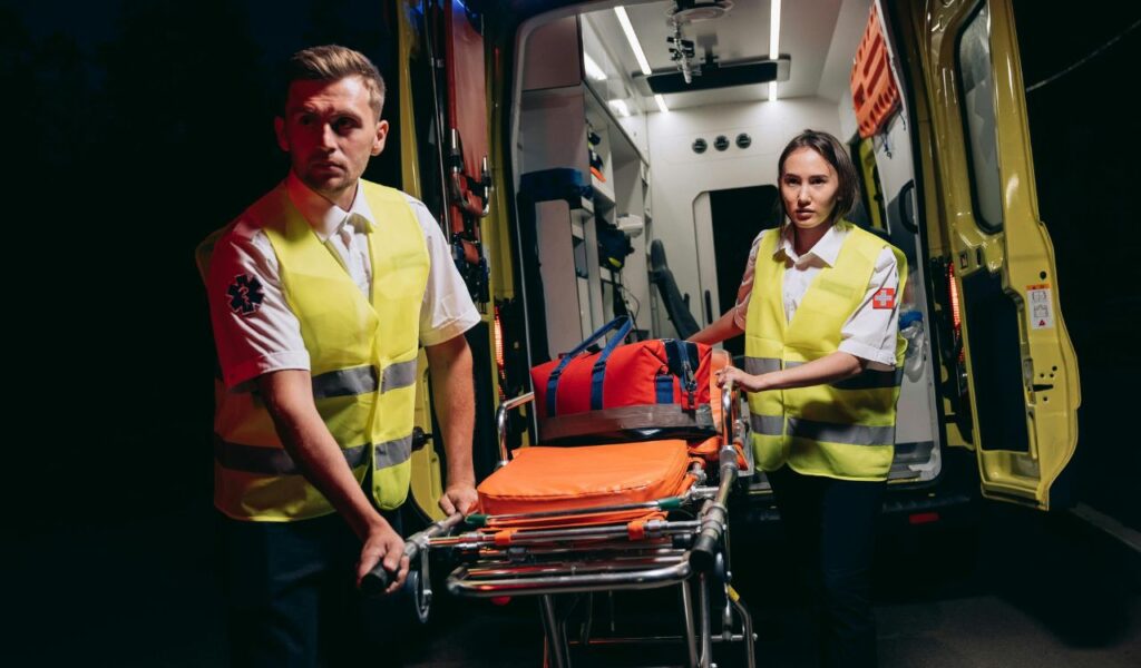 Two emergency medical responders wheel a stretcher toward an ambulance at night, symbolizing urgent care and the challenges of reaching help quickly in remote locations.