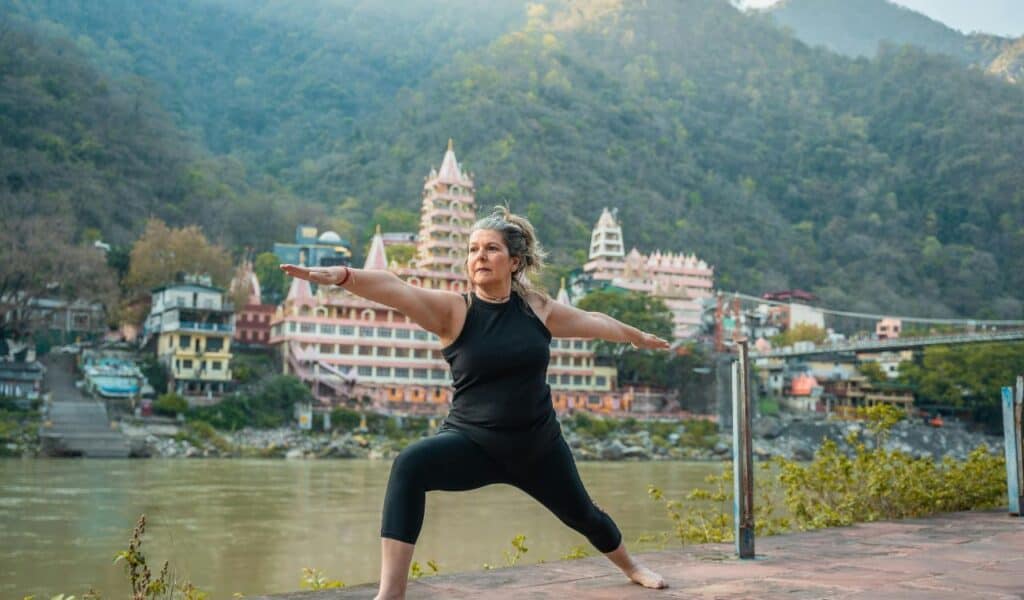  Woman practicing yoga in warrior pose beside the Ganges River, with colorful temples and buildings of Rishikesh and forested hills in the background.