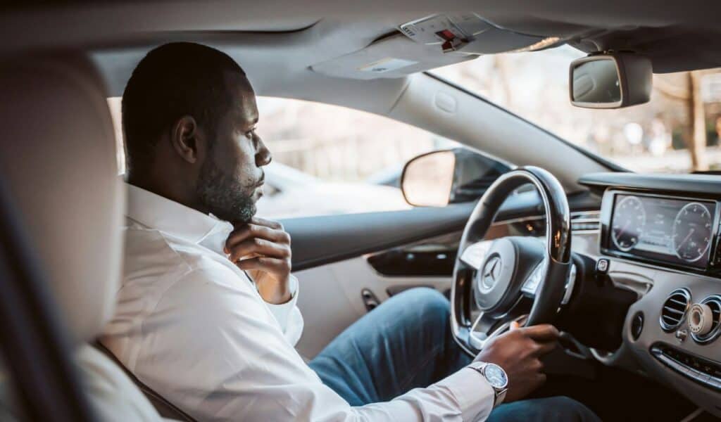 : A man wearing a white shirt sits in the driver's seat of a modern car, adjusting his collar while holding the steering wheel, suggesting he is preparing to drive.