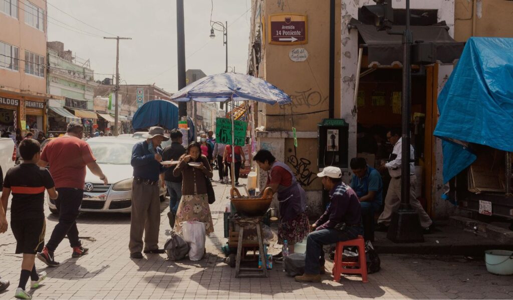 Street food vendor serving traditional Mexican dishes to locals under a blue umbrella in a busy market area in Mexico.