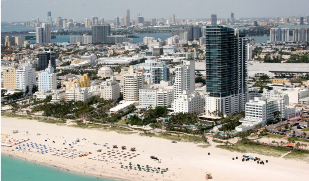 Aerial view of Miami Beach with white sand, colorful umbrellas, and high-rise buildings near the shore.