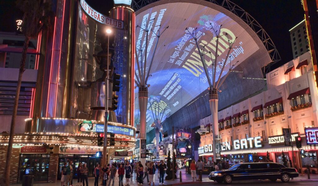 A bustling Las Vegas street at night, showcasing bright neon signs, a large illuminated overhead screen, and people walking along Fremont Street with a limousine passing by.