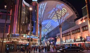 A bustling Las Vegas street at night, showcasing bright neon signs, a large illuminated overhead screen, and people walking along Fremont Street with a limousine passing by.