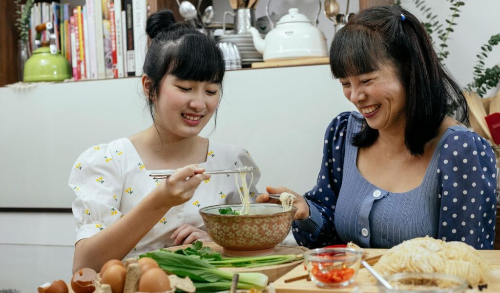 Two women smiling and using chopsticks to eat noodles at a table with fresh vegetables, eggs, and condiments, showcasing traditional dining etiquette and shared enjoyment of food