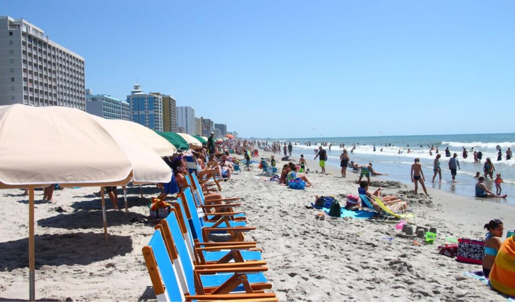 Crowded Myrtle Beach shoreline with sunbathers, umbrellas, and oceanfront hotels under a clear blue sky.