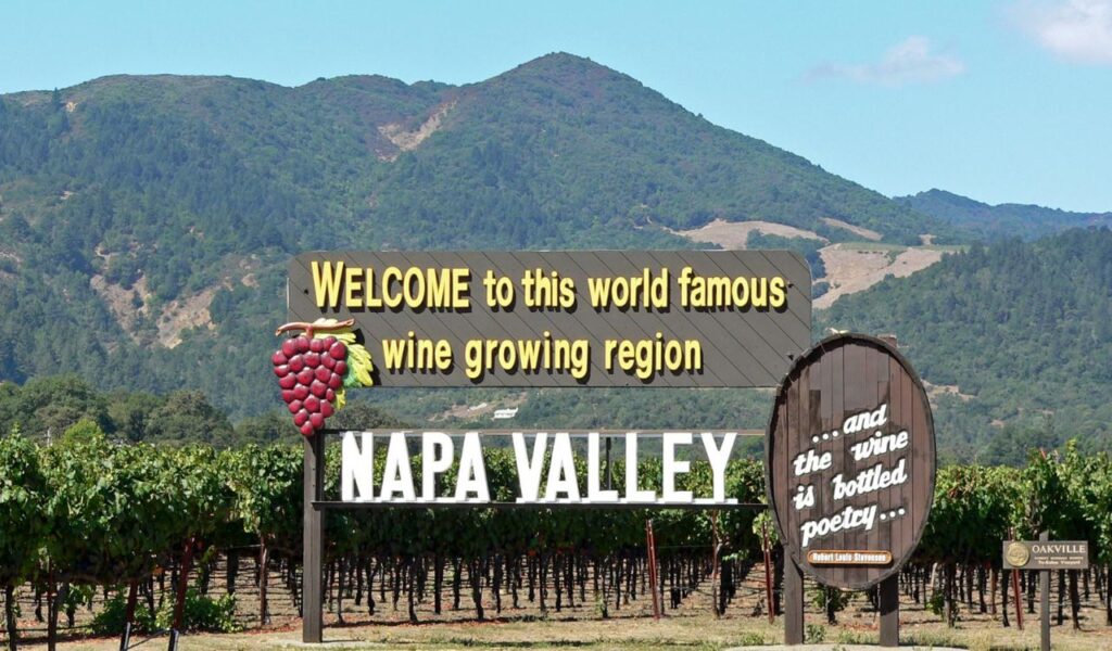 Welcome sign for Napa Valley, California, set against green vineyards and tree-covered hills in the background.
