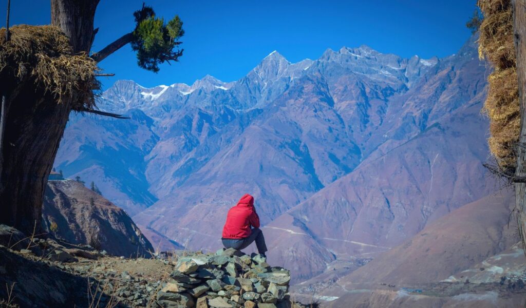 Solo traveler in red jacket sitting on rock, admiring snow-capped mountains in Nepal.